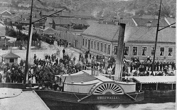 Trängsel runt hjulångaren ”Uddewalla” år 1865. Till vänster syns den minimala biljettkiosken. Vändbassängen, som anlades delvis för ”Uddewallas” skull, ligger på Västerbrons nuvarande plats. Ångfartyget ”Uddewalla” var en praktsensation när det år 1853 levererades från Motala verkstad. Med sina 60 hästkrafter var det Sveriges snabbaste fartyg. Sträckan mellan Göteborg och Uddevalla – som med tidigare förbindelser tagit dagar eller rentav veckor – tog plötsligt två timmar. Det ansågs av samtiden var chockartat snabbt. Det betraktades också som vådligt och dödsföraktande att gå i öppen sjö. När bilden togs var dock ”Uddewalla” redan på väg att bli omodernt. Inom några år skulle hjulångarna ha slagits ut av propellerfartyg. Fotografiet togs troligen i mitten av 1860-talet av Maria Lundbäck – en av Uddevallas första professionella fotografer. Hon använde troligen den då populära våtplåtsmetoden som gjorde det möjligt att ta detaljerade bilder, men som krävde framkallning inom 10-15 minuter. Några människor har rört sig under exponeringen och kan följaktligen anas som grå och spöklika dubbelfigurer på kajen.