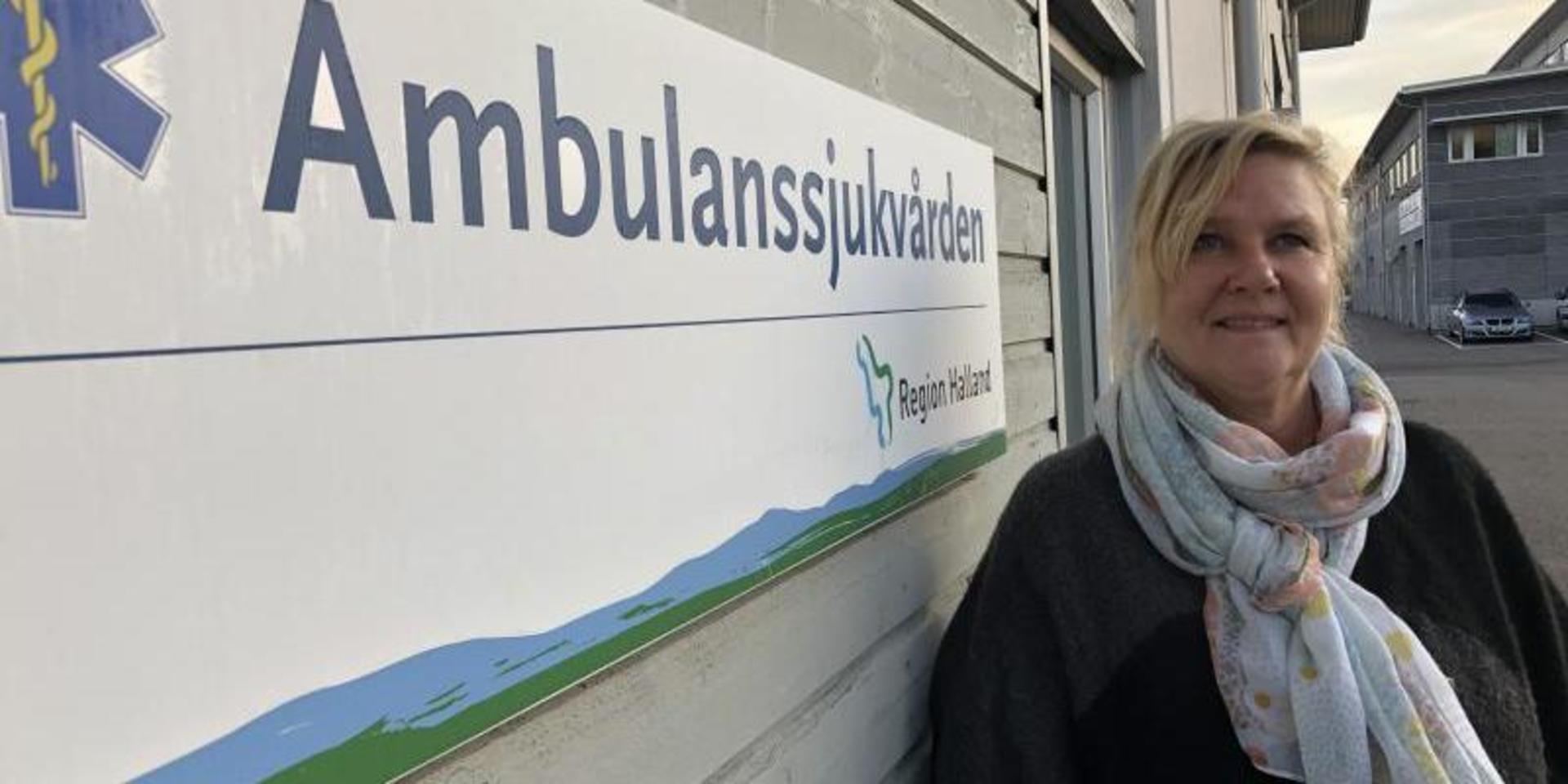 Kvinna i ljus sjal med ljusa färger står framför en grå vägg med ordet Ambulanssjukvården. 